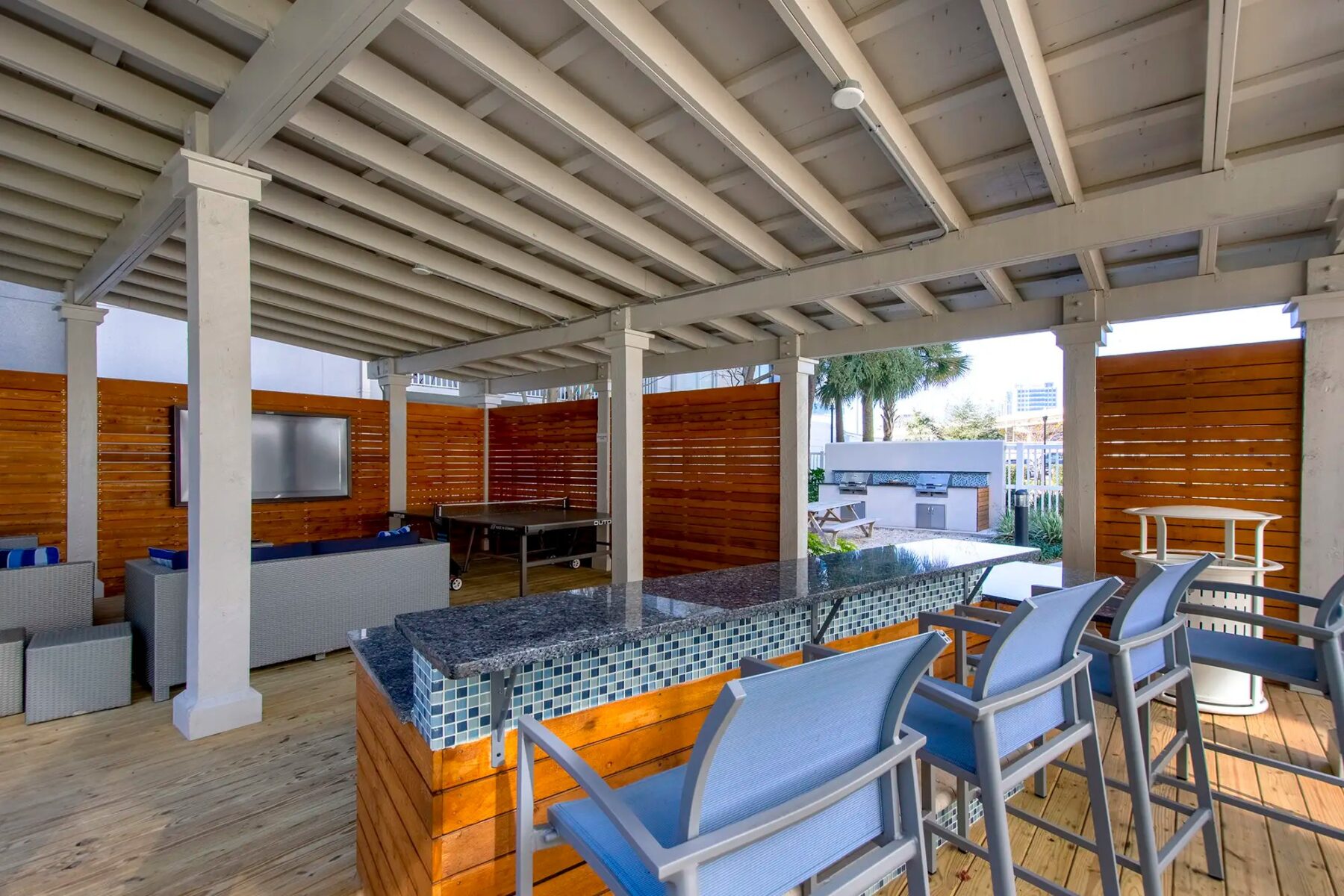 Covered outdoor area with bar and barstools. pingpong table, couches and tv mounted on the wall.