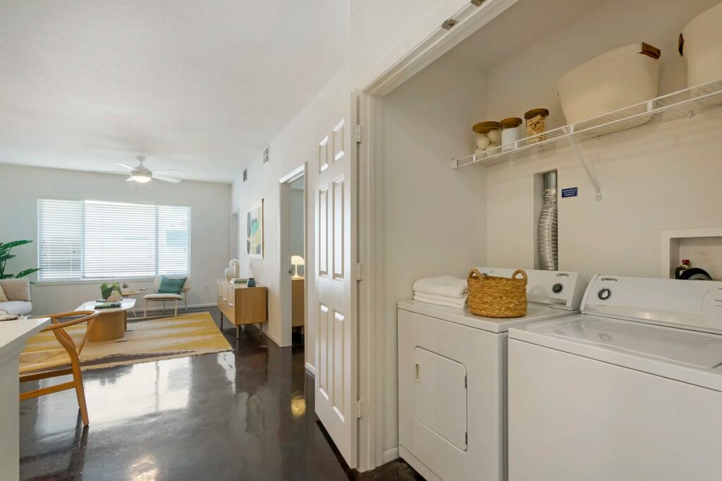 Open laundry room with washer and drier and built in shelf above. With view of the living room to the left.